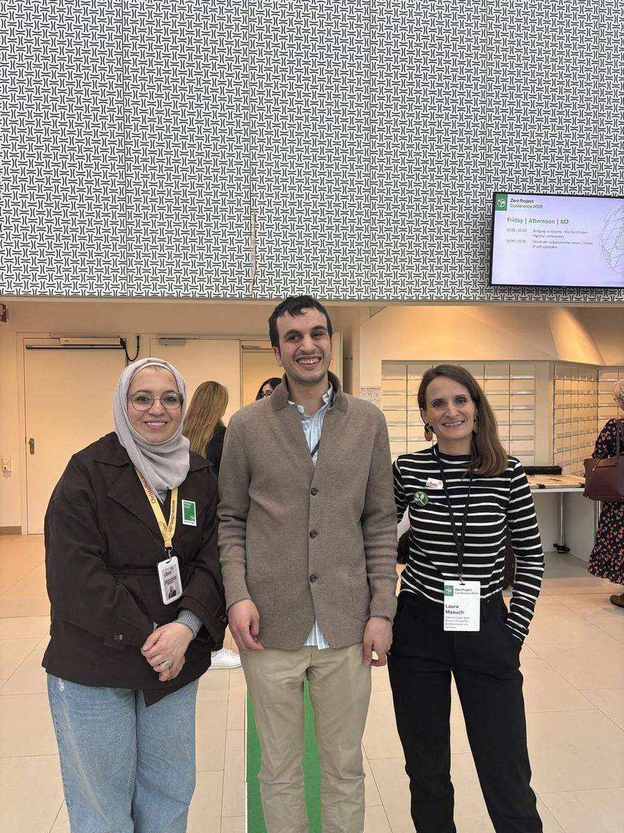 Hasan with two colleagues at a conference session, all wearing conference badges, with a presentation screen visible in the background