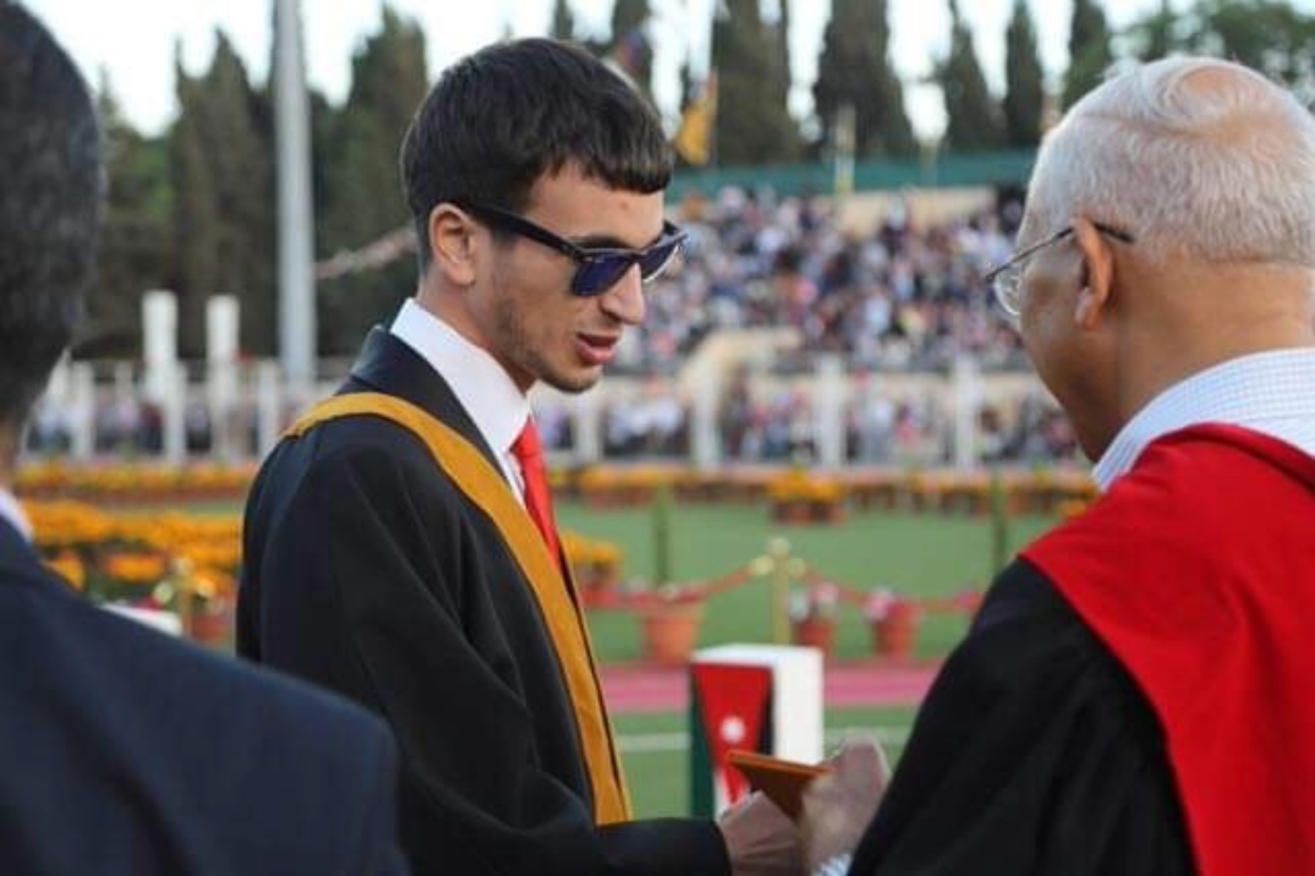 Hasan, wearing graduation robes and sunglasses, celebrates his bachelor's graduation outdoors by shaking hands with a distinguished guest