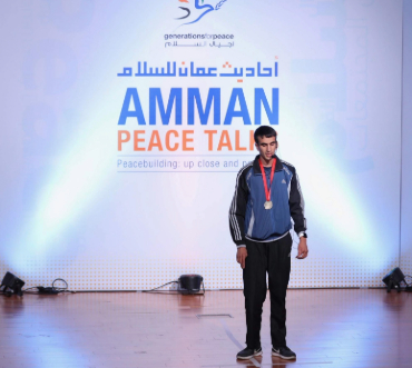 Hasan stands confidently on stage at the Amman Peace Talks event, highlighted by blue lights and the event banner behind him