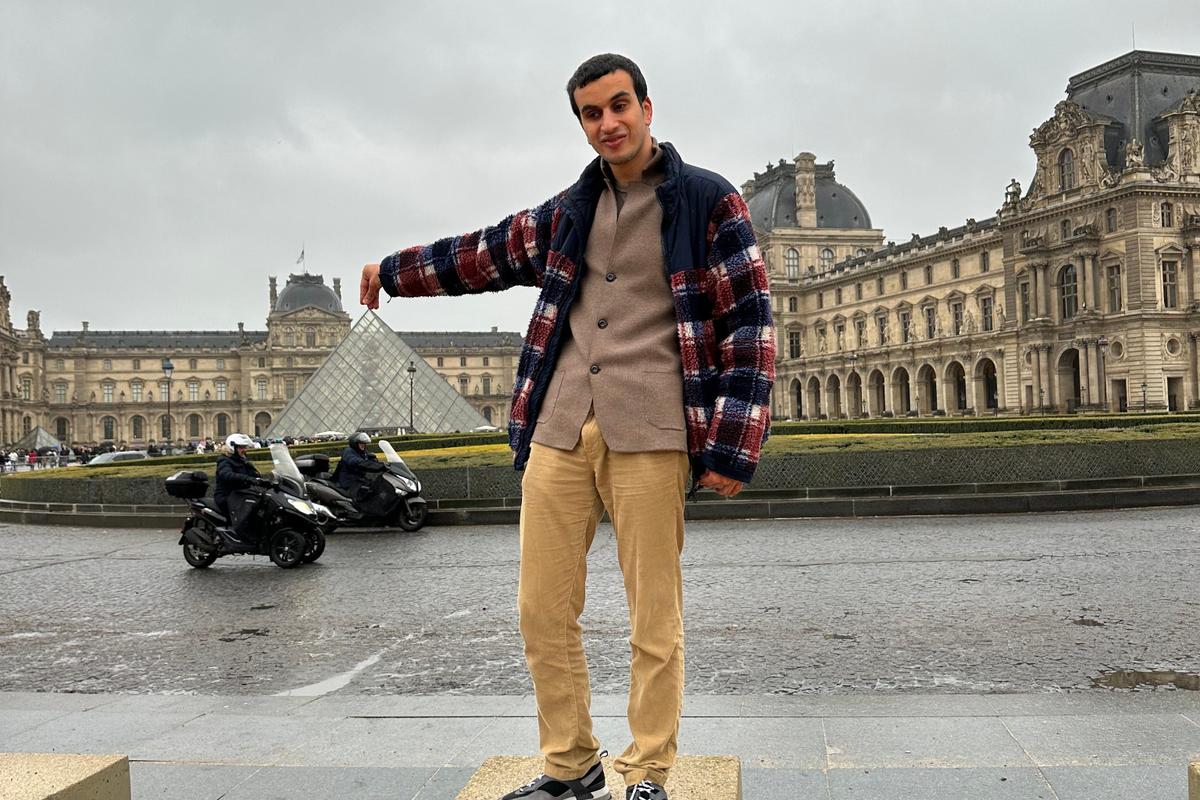 Hasan standing in front of the Louvre Museum in Paris, playfully posing as if he is touching the top of the glass pyramid with his hand, on a cloudy day