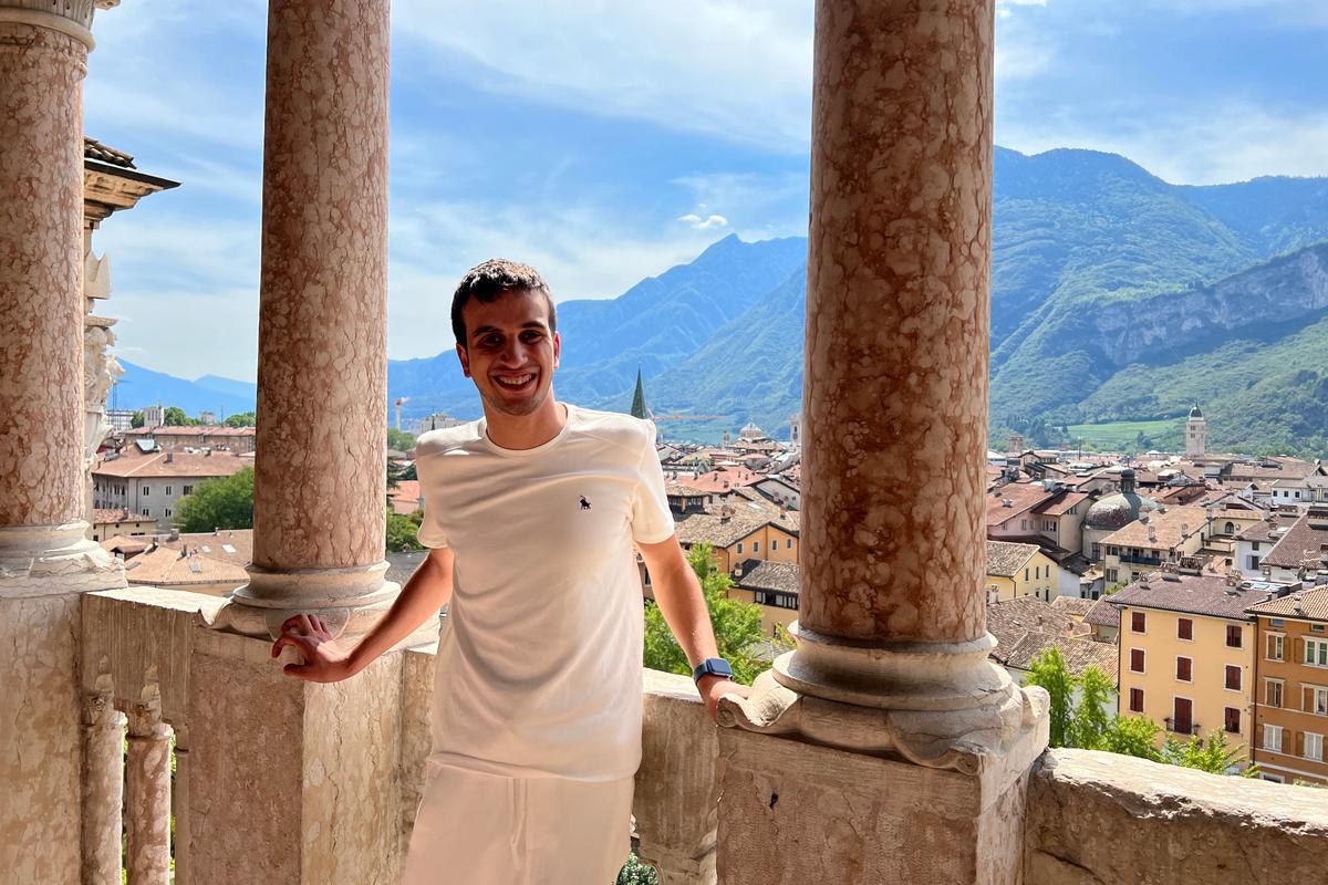 Hasan leaning against a stone balcony with columns, overlooking a picturesque town with mountains under a bright blue sky